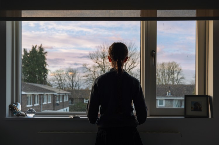 Teenage girl looking out a bedroom window at dusk.